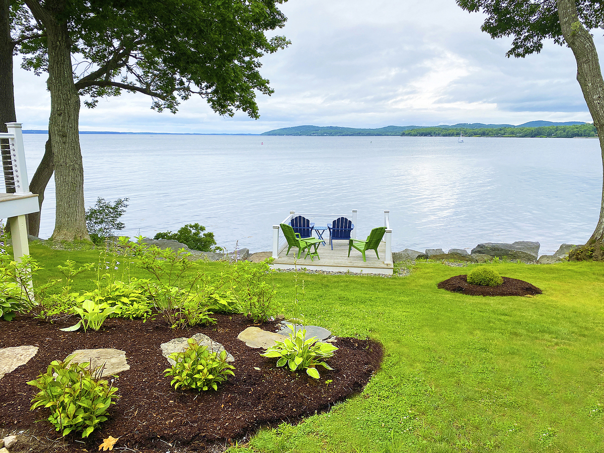 Oceanfront property with green grass, adirondack chairs, trees, plants behind it